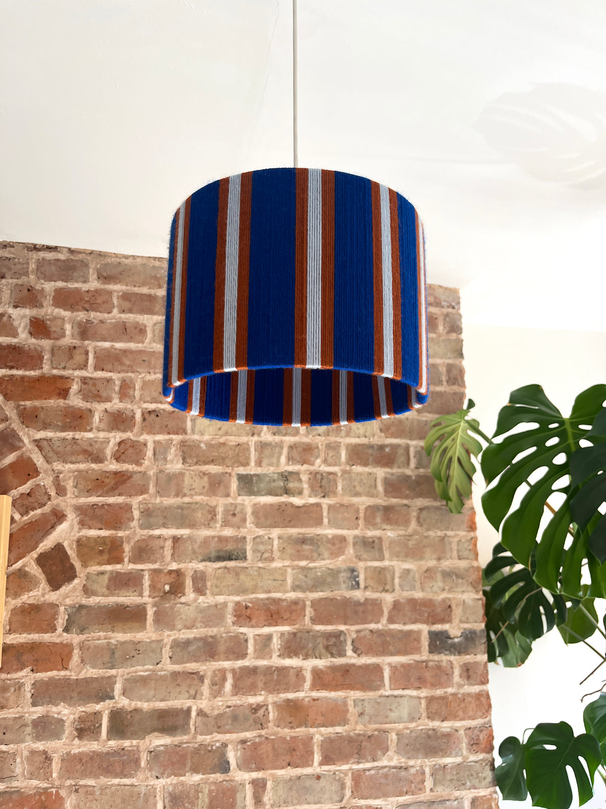 A drum-shaped A Slice Shades yarn drum lampshade with vertical blue, orange, and white stripes hanging against a brick wall background.