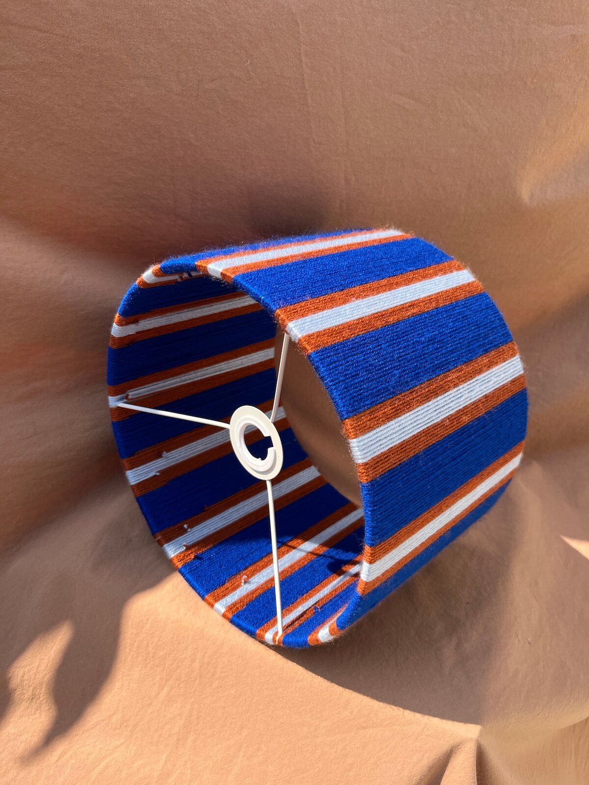 A Slice Shades yarn drum lampshade  with vertical blue, orange, and white stripes with a striped pattern of royal blue, orange, and white, shown on a draped tan fabric background.