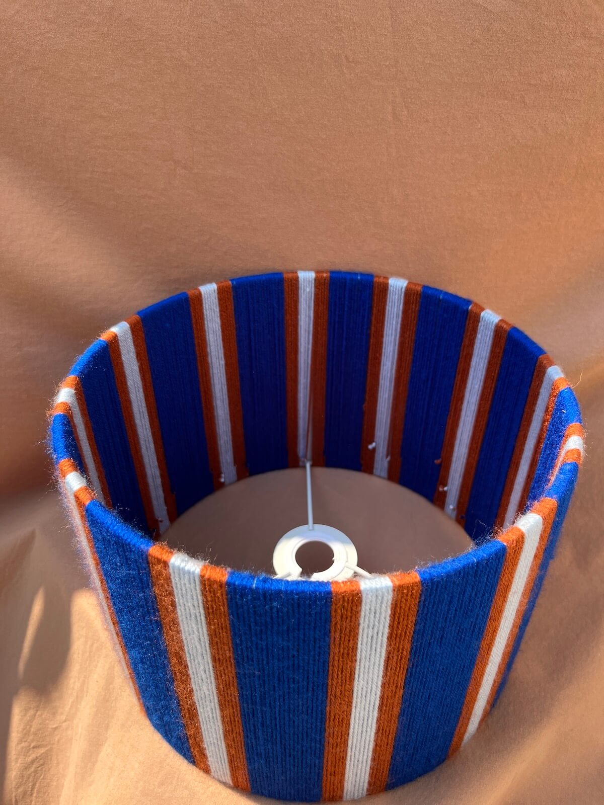 A Slice Shades yarn drum lampshade  with vertical blue, orange, and white stripes with vertical stripes in royal blue, orange, and white, photographed against a warm tan fabric background.
