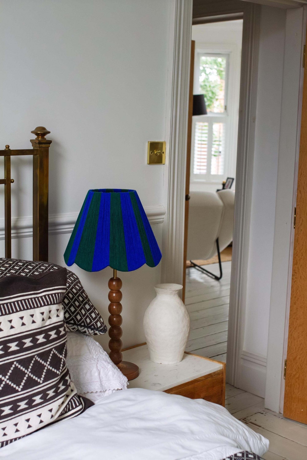 A bedroom scene featuring a table lamp with a slice shades yarn blue and green striped fabric shade and a turned wooden base, styled on a bedside table next to a white ceramic vase.