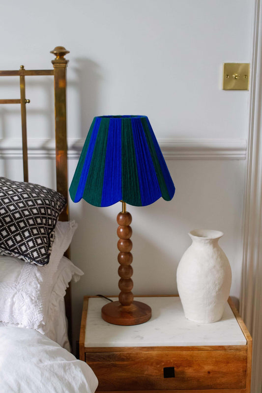 A scallop table lamp with a vibrant blue and green striped yarn lampshade and a turned wood base, styled on a bedside table next to a bed and a white ceramic vase.