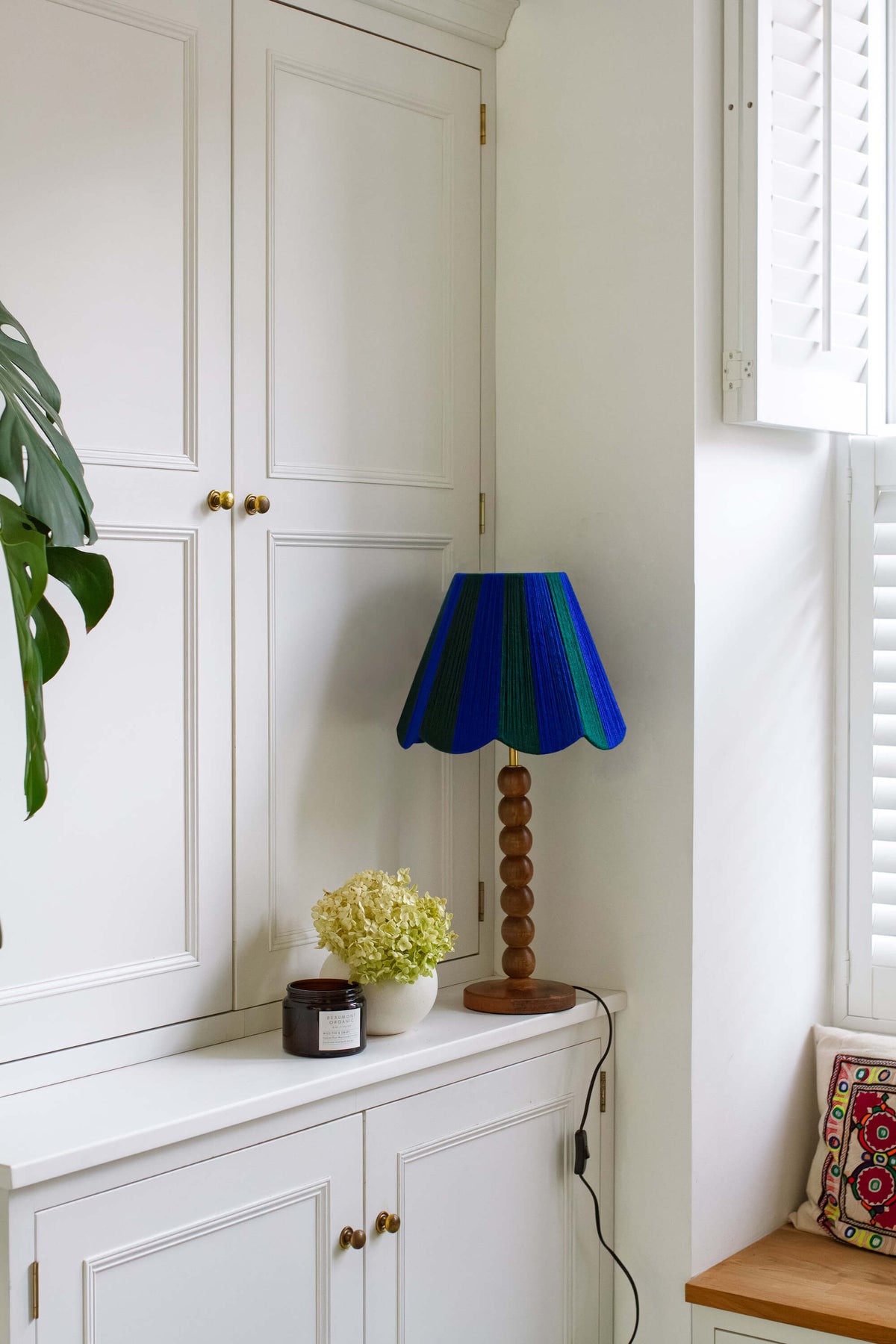 A decorative table lamp with a slice shades yarn blue and green striped lampshade and a turned wooden base, styled on a white sideboard in a bright room.
