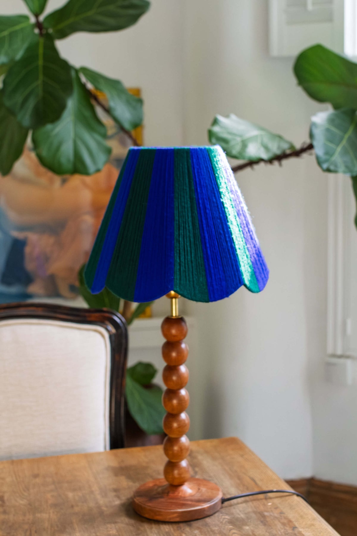 A table lamp with a a slice shades yarn  lampshade in alternating vertical stripes of royal blue and emerald green, supported by a polished wooden bead spindle base on a wooden table.