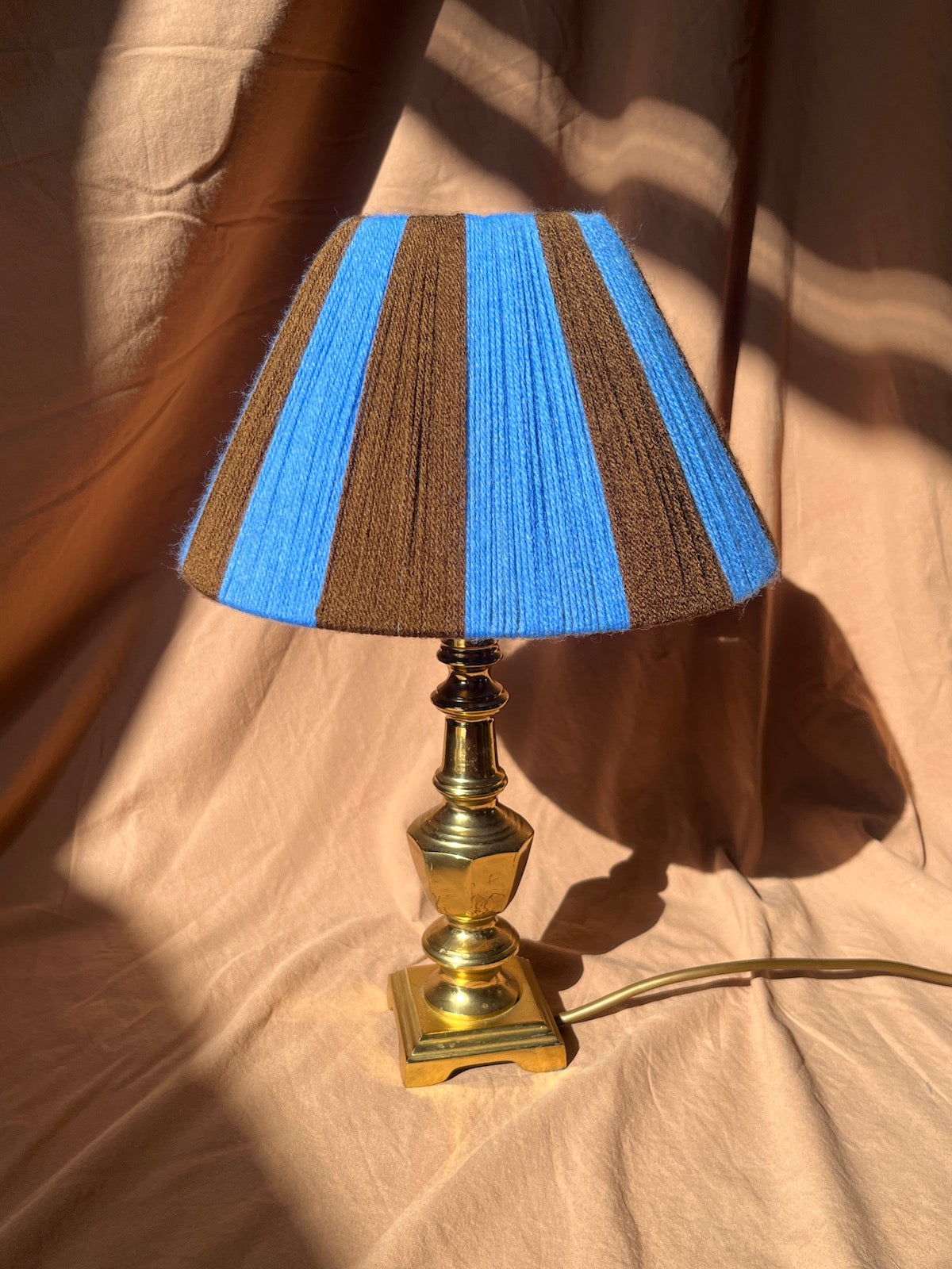 A table lamp with a Slice Shade featuring alternating vertical stripes of bright blue and brown, mounted on a polished brass-finished base with a cord extending to the right, set against a draped beige fabric backdrop with strong sunlight and shadows.