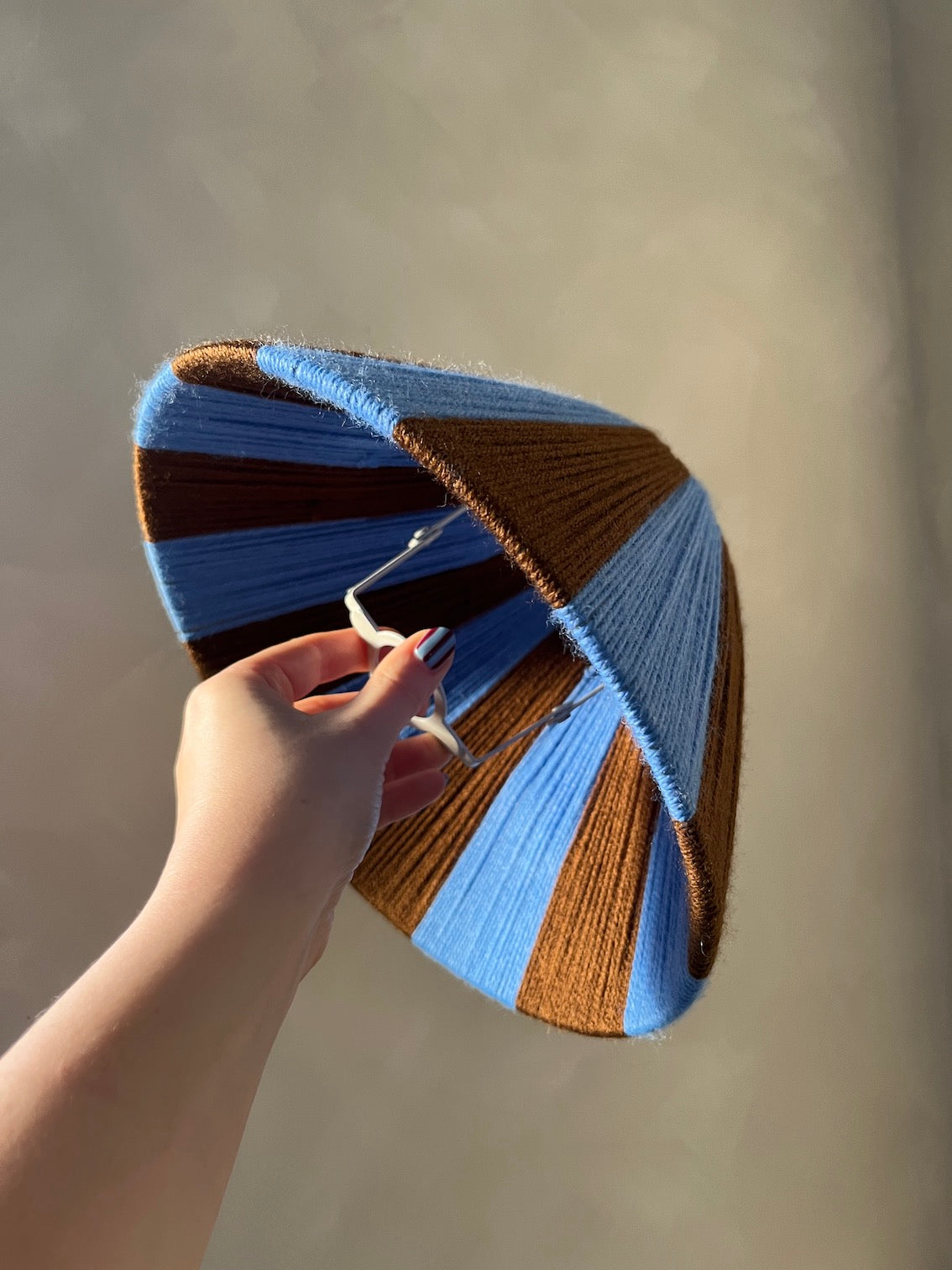 A hand holding a Slice Shades handwoven striped lampshade with alternating blue and brown stripes bands against a plain beige background.