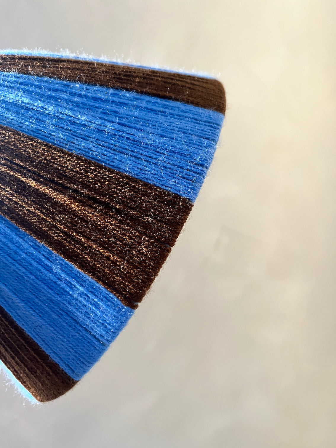 A close-up of a textured Slice Shade lampshade,  featuring stripes of blue and brown yarn on a light beige background.
