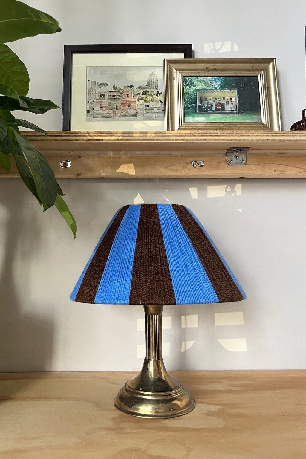 A table lamp with a blue and dark brown striped yarn Slice Shades shade and a brass-colored metal base, placed on a light wood surface in front of a white wall with a wooden shelf and framed pictures behind it.