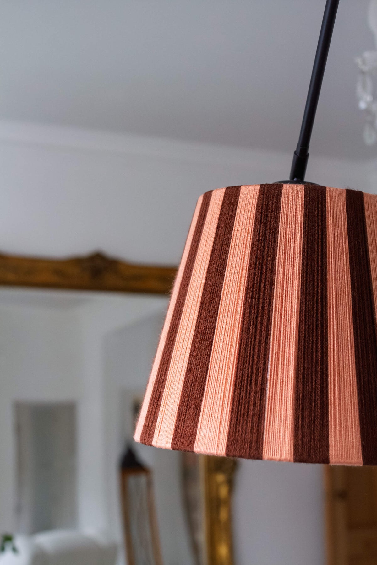 A close-up of a slice shades handwoven yarn lampshade with vertical stripes in cocoa and plaster pink, hanging from a black cord in an indoor room setting.