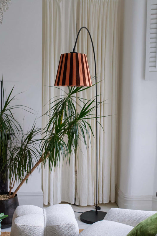 A black lamp stand with a slice shades handwoven yarn lampshade with vertical stripes in cocoa and plaster pink, hanging from a black cord in an indoor room setting.