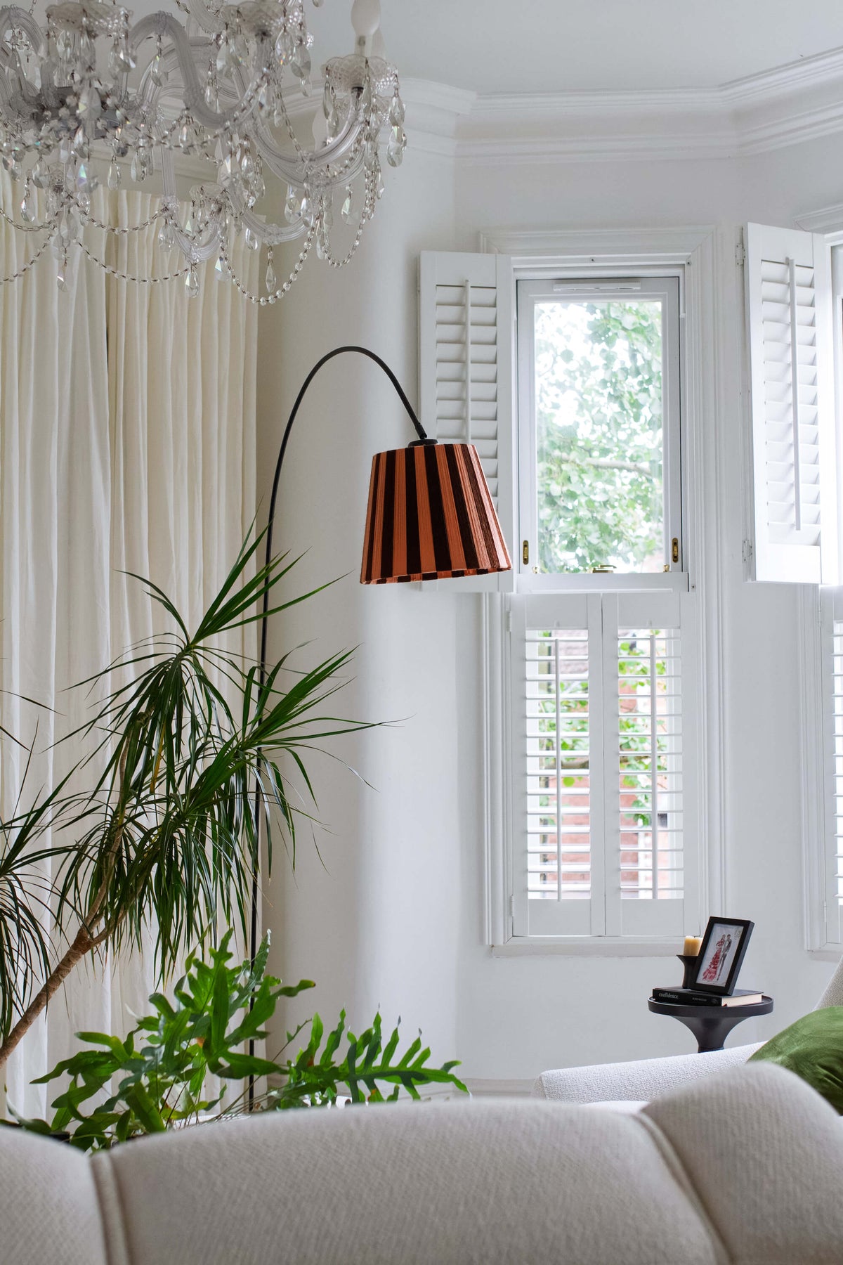 A modern floor lamp with a black metal frame and slice shades yarn striped lampshade in cocoa and plaster pink, positioned in a bright living room next to a window with white shutters.