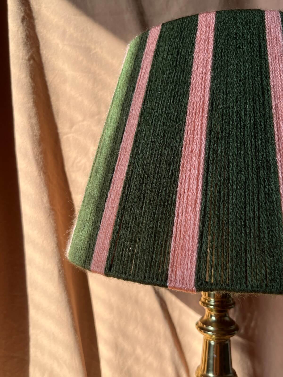 A close-up of a striped string yarn lampshade with vertical bands of dark green and pink stripes, mounted on a brass lamp base against a draped beige fabric background.