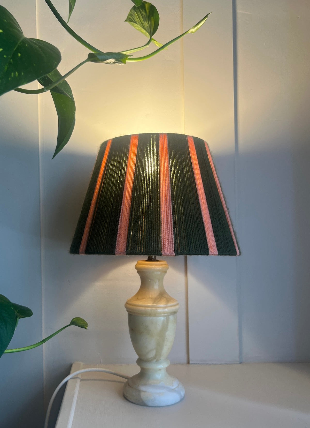 A table lamp with a string lamp shade featuring vertical stripes of green and pink mounted on a polished brass base and set on a light wood surface against a panelled backdrop.