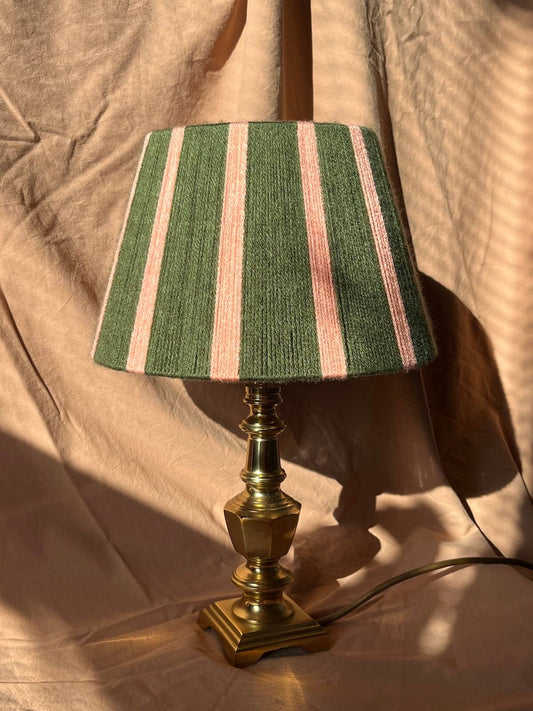 A table lamp with a green,and pink striped string shade and a brass-colored metal base, set against a draped beige fabric background.