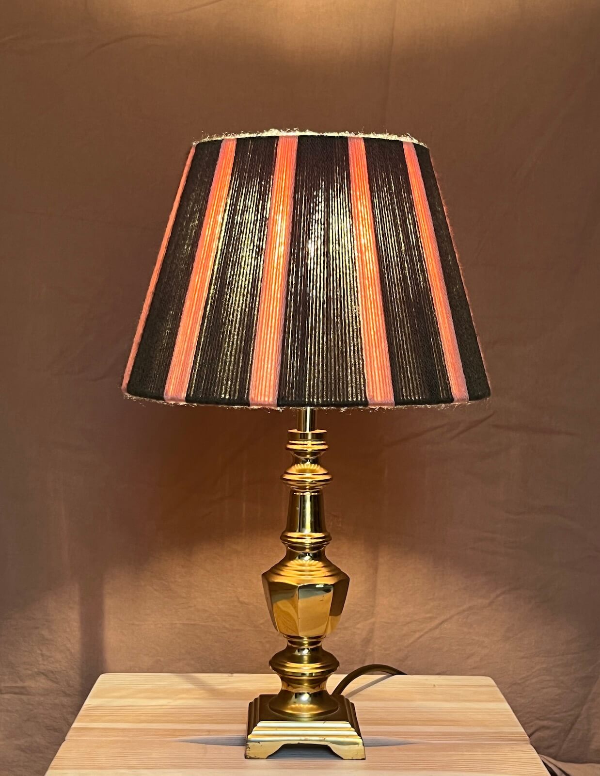 A table lamp with a string lamp shade featuring vertical stripes of green and pink mounted on a polished brass base and set on a light wood surface against a brown backdrop.
