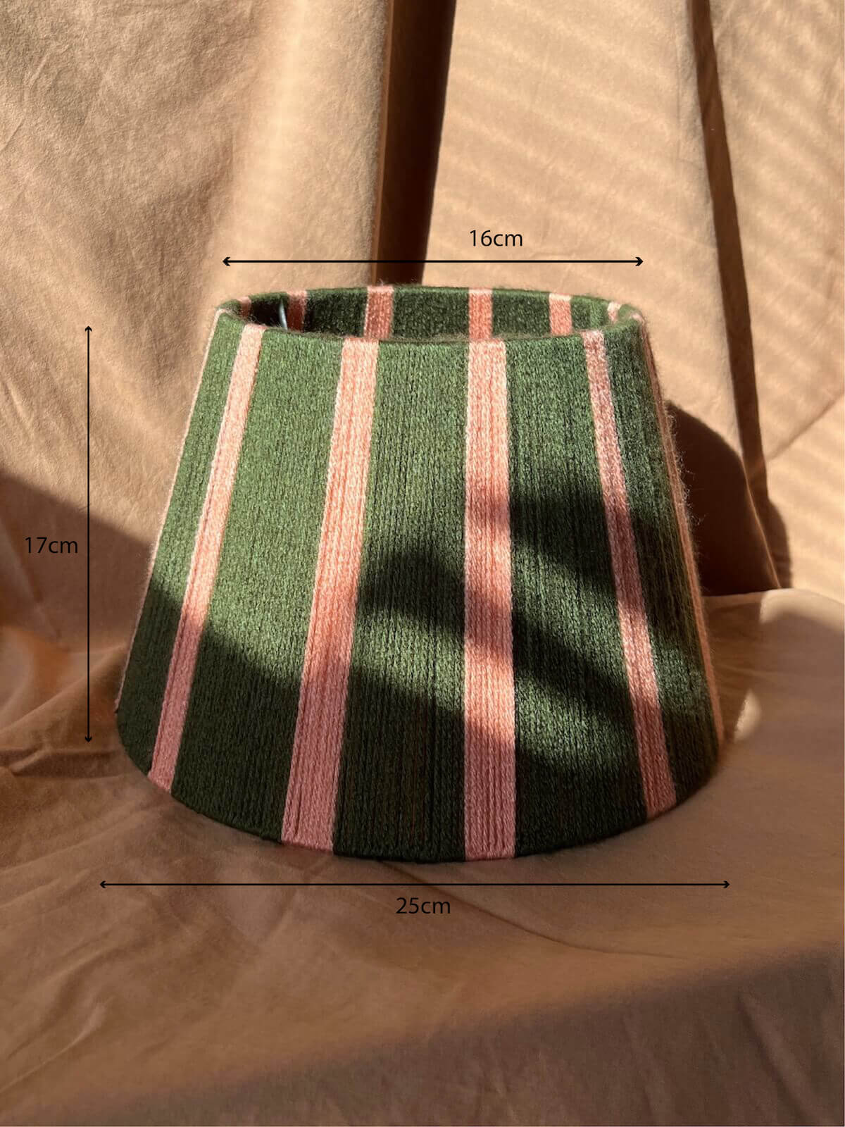 A handwoven string lampshade with vertical stripes in dark green and light pink, resting on a beige fabric backdrop with warm sunlight and shadows.