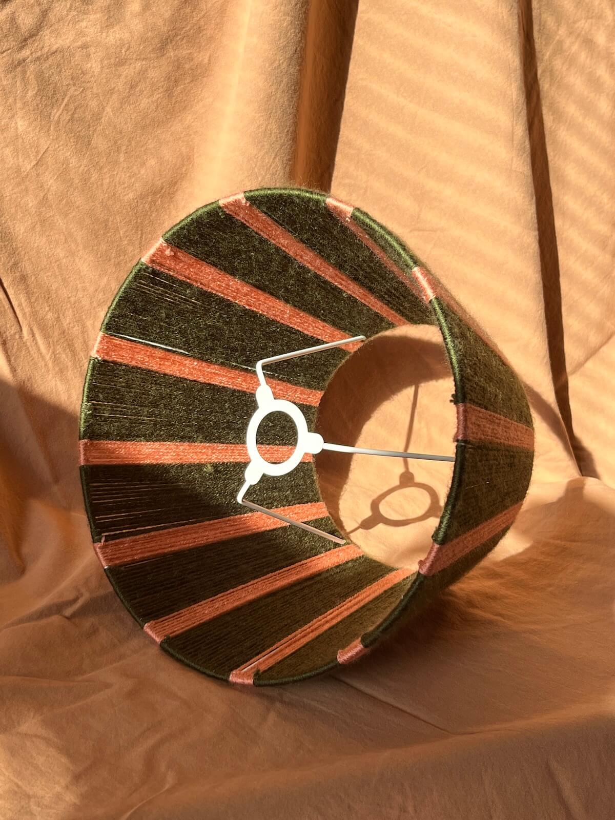 A conical lampshade with alternating dark green and pink stripes handwoven in yarn shown on a draped beige fabric background.