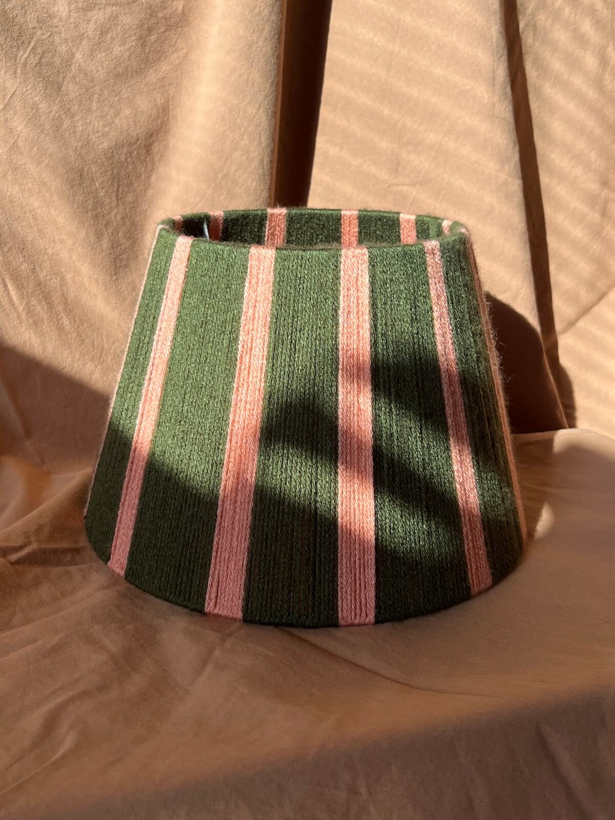 A handwoven string lampshade with vertical stripes in dark green and light pink, resting on a beige fabric backdrop with warm sunlight and shadows.