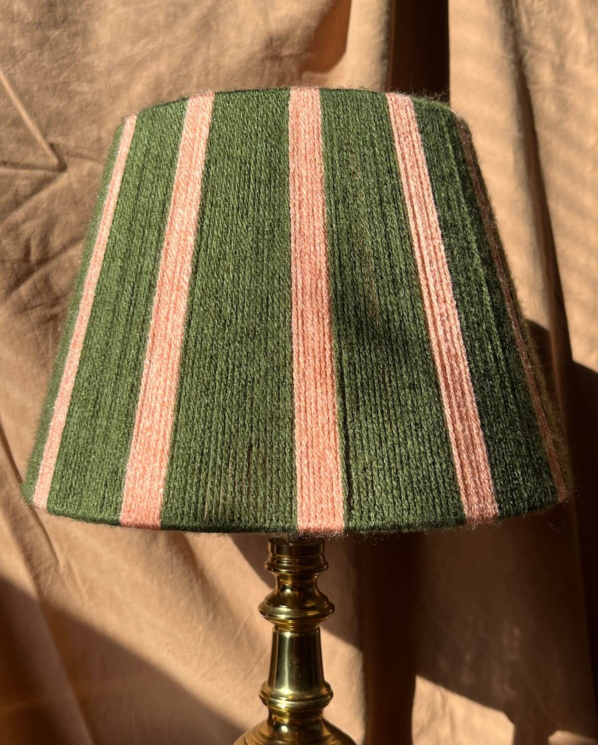 A table lamp with handwoven string shade featuring vertical stripes of dark green and light pink in yarn mounted on a polished brass base against a draped beige fabric backdrop.