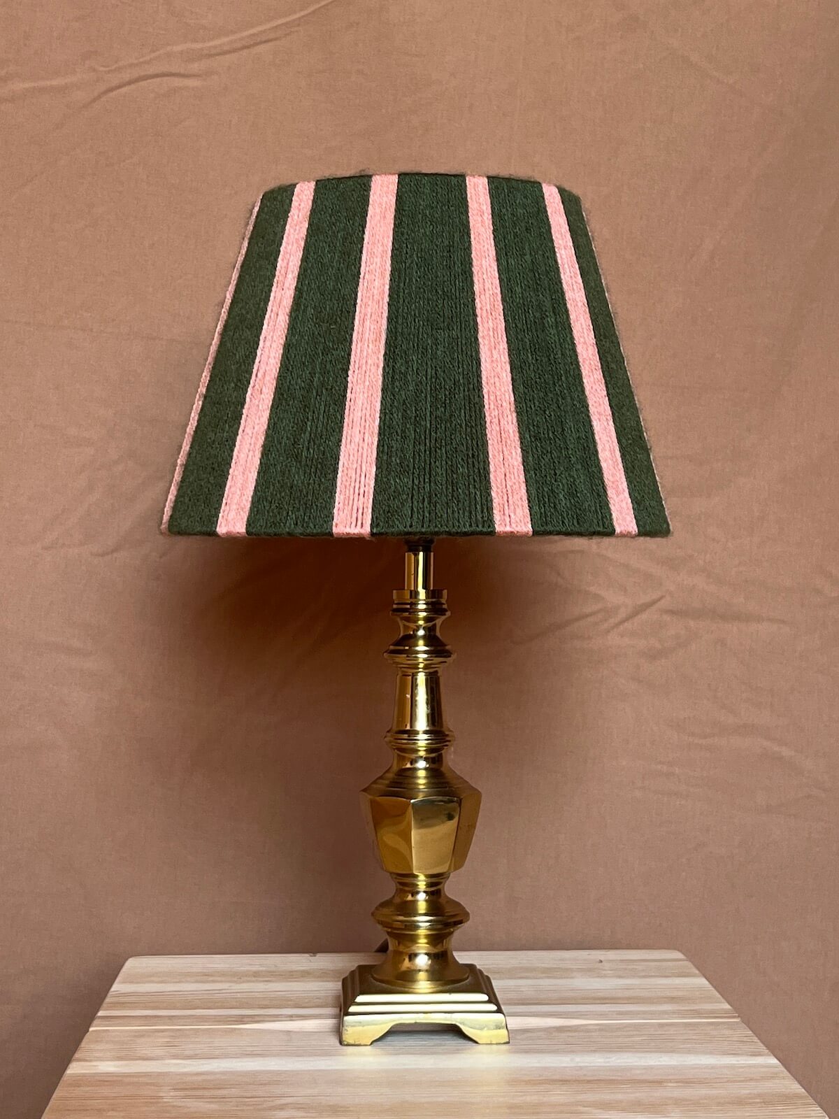 A table lamp with a dark green and pink striped string lamp shade and a polished brass base, set on a light wood surface against a brown backdrop.
