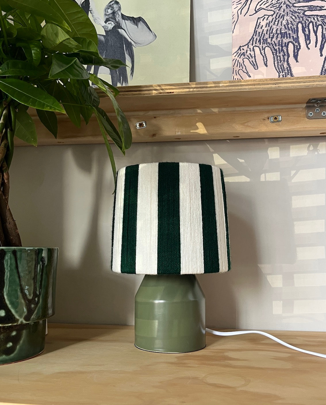 A Slice Shades table lamp with a green and white vertically striped yarn drum shade and an olive green ceramic base, sitting on a light wood shelf next to a potted plant.