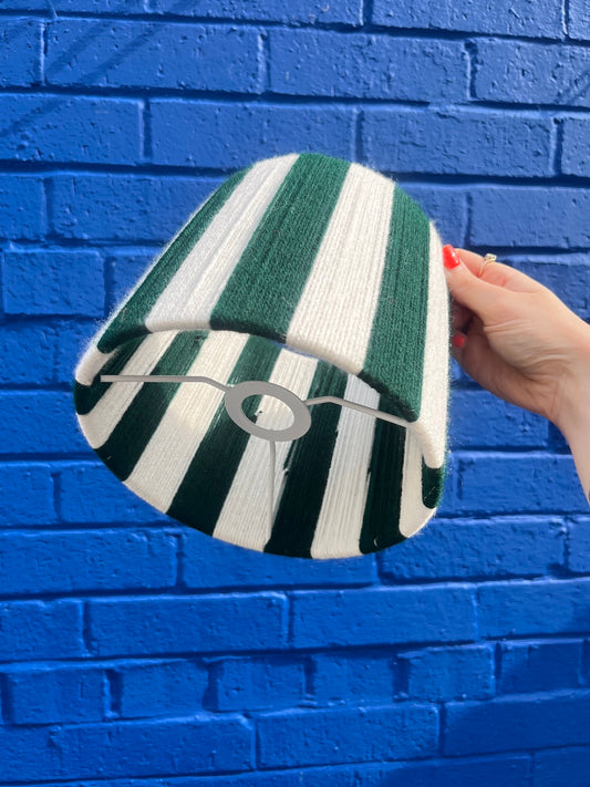 A hand holding a Slice Shades lampshade with vertical green and white stripes, set against a bright blue brick wall.