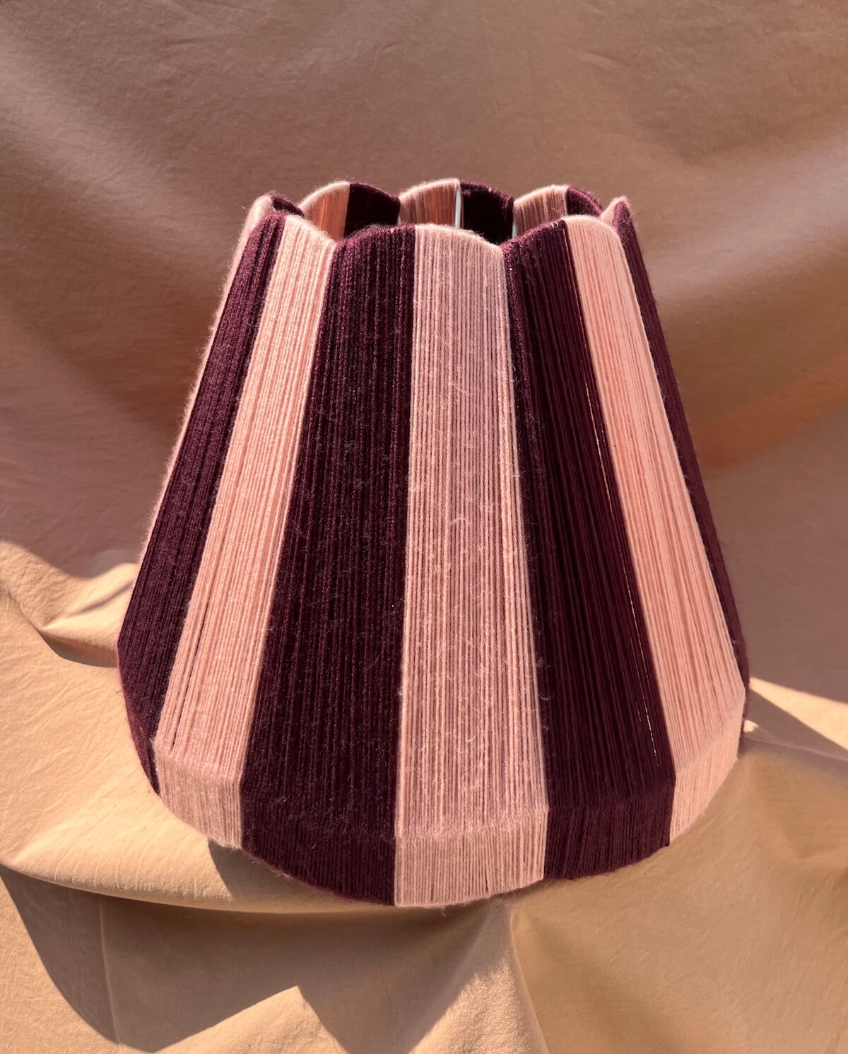 A Slice Shades large scallop yarn lampshade with alternating vertical stripes of deep plum purple and dusty pink, set against a draped beige fabric backdrop.