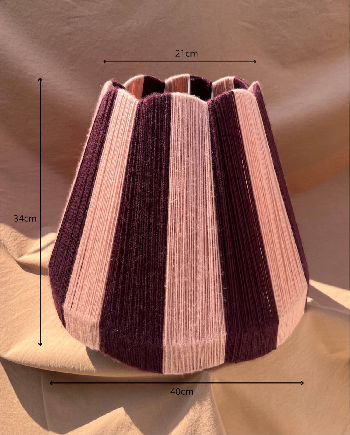 A Slice Shades large scallop yarn lampshade, featuring alternating stripes of deep plum purple and light blush pink, set against a draped beige fabric background with measurments.