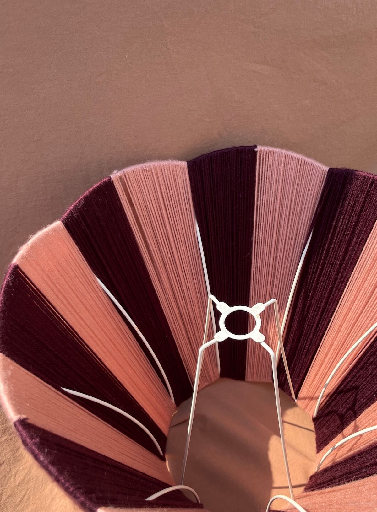 A close-up of a A Slice Shades large scallop yarn lampshade in alternating shades of dusty pink, mauve, and deep burgundy, supported by a white wire frame.
