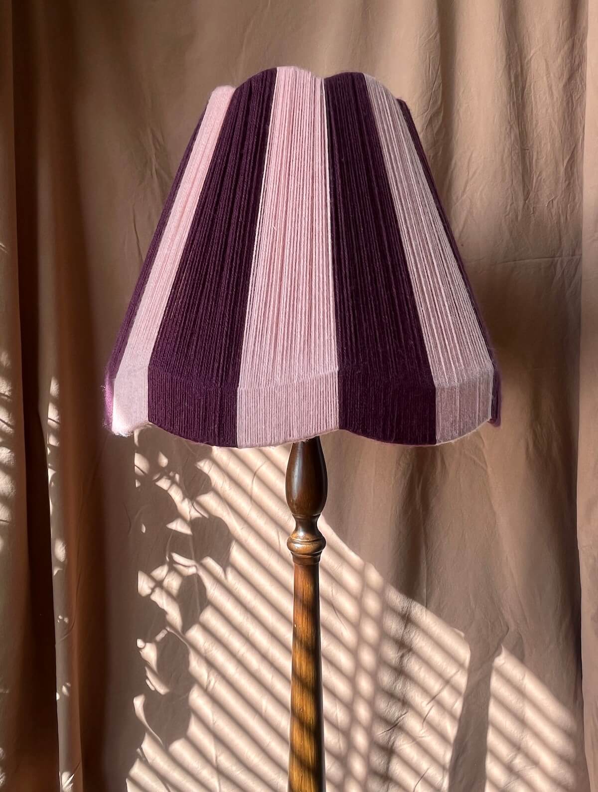 A Slice Shades large scallop yarn lampshade in alternating vertical stripes of deep purple and light lavender, mounted on a dark brown wooden pole, set against a draped beige fabric backdrop with window shadow patterns.