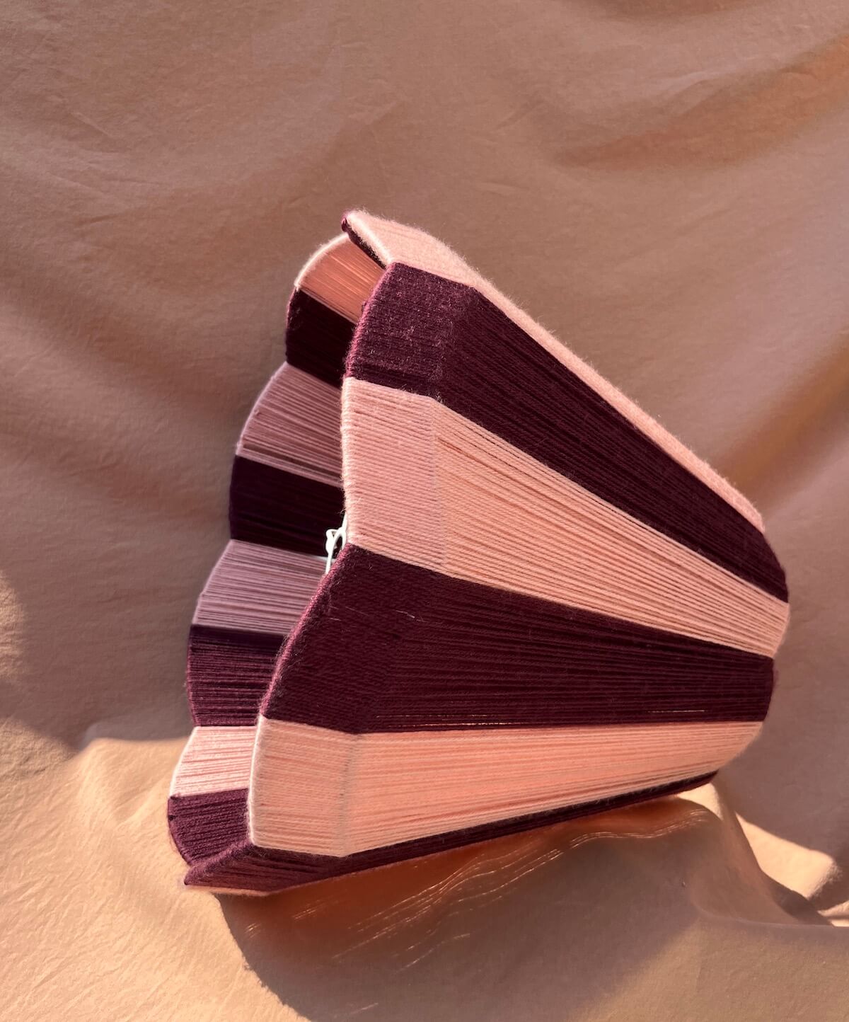 A Slice Shades large scallop yarn lampshade, featuring alternating stripes of deep plum purple and light blush pink, set against a draped beige fabric background.