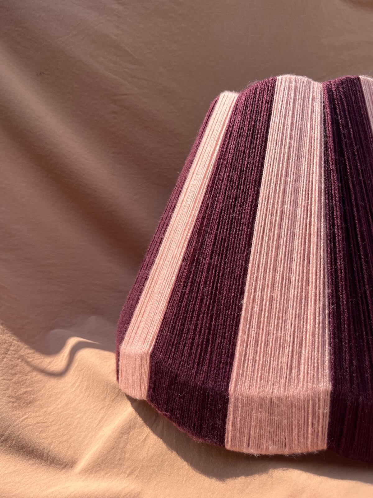 A close-up of a Slice Shades large scallop yarn lampshade alternating vertical stripes in deep plum purple, dusty pink, and pale blush, set against a draped beige fabric background.