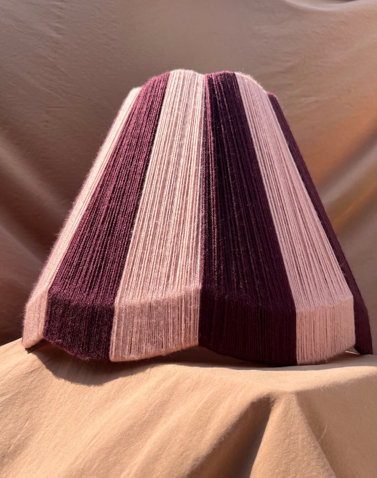 A Slice Shades large scallop yarn lampshade, featuring alternating stripes of deep plum purple and light blush pink, set against a draped beige fabric background.