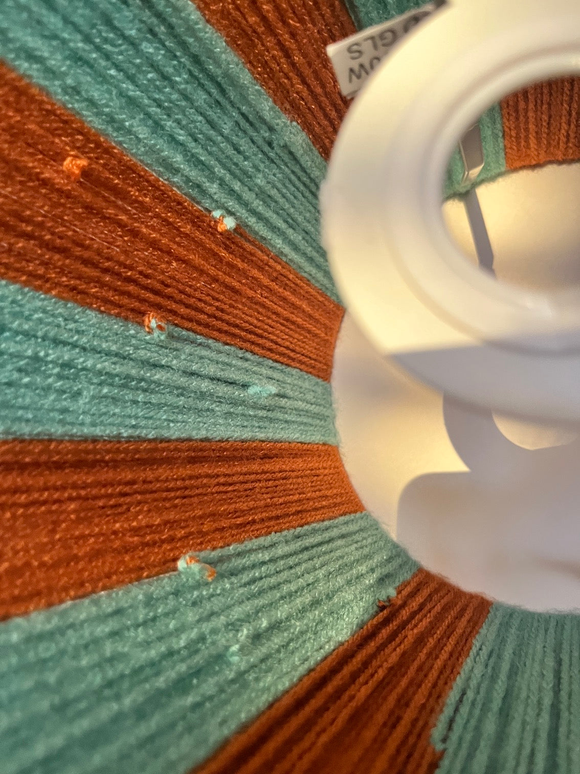 Close-up of a spiral CFL light bulb inside a lamp socket, showing textured orange and teal striped fabric or mesh around the bulb.