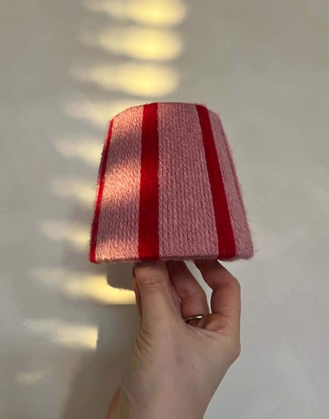 A hand holding a small Slice Shades yarn lampshade with vertical pink and red stripes against a light wall with soft shadow patterns.