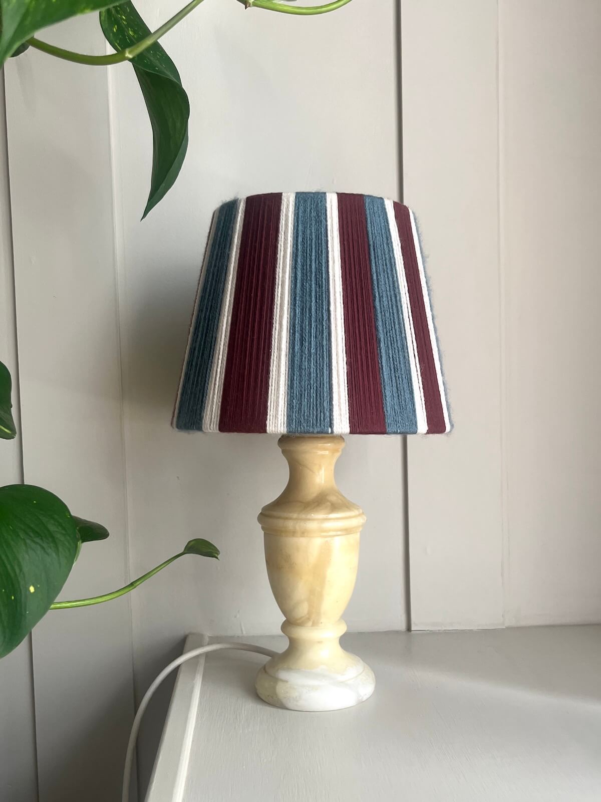 A table lamp with a Slice Shades yarn lampshade featuring vertical stripes of burgundy, slate blue, and white, set on a light-colorued surface against a white panelled wall with green plant leaves in the foreground.