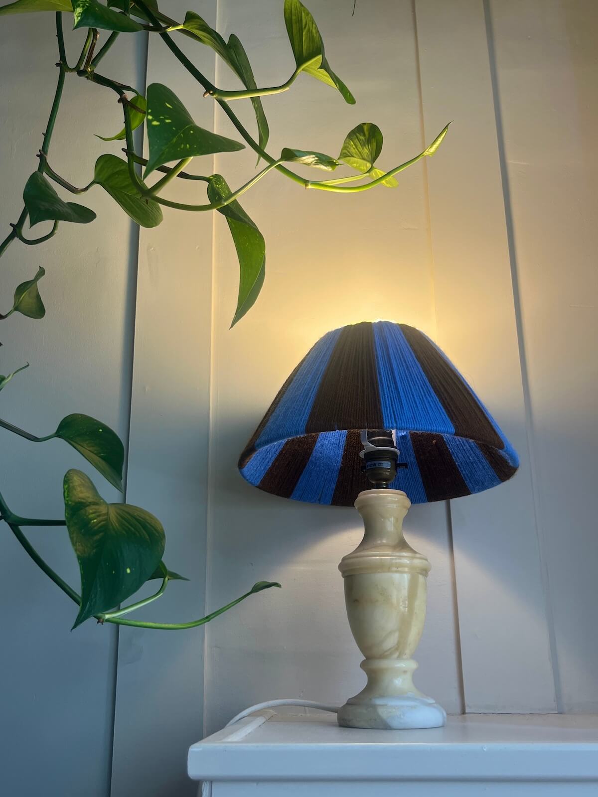 A table lamp with a Slice Shades blue and brown striped yarn shade and a metallic gold base, shown on a wooden surface glowing.