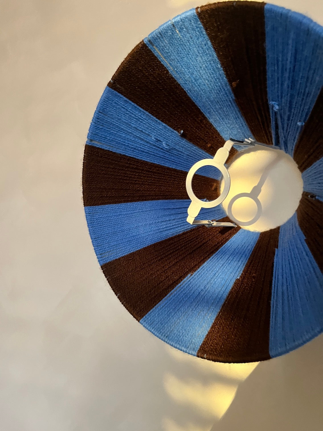 A close up of the inside of a Slice Shades lampshade with alternating stripes of medium blue and dark brown, featuring a white metal socket at the center, shown against a plain light background.