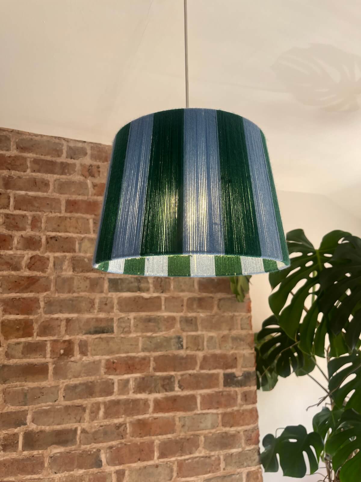 A Slice Shades handmade yarn drum lampshade glowing in front of an exposed brick wall.