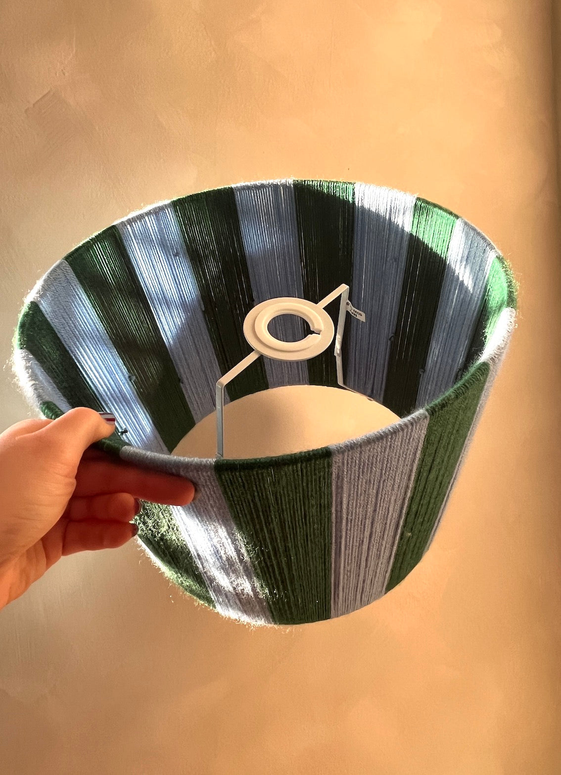 A hand holding a Slice Shades handmade yarn drum lampshademade from woven striped yarn in shades of green, blue, and gray, against a plain beige wall.