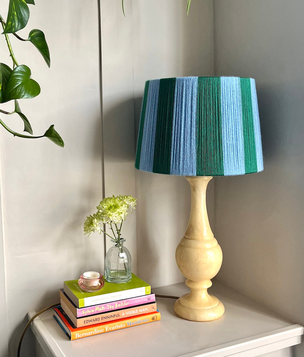 A  Slice Shades handmade yarn drum lampshade in vertical stripes of blue and green, set on a white side table next to a stack of colourful books and a small glass vase with white flowers.