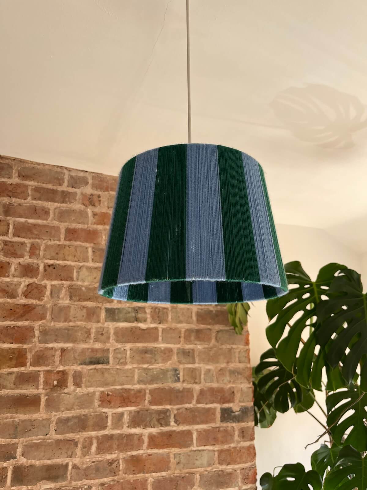 A Slice Shades handmade yarn drum lampshade glowing in front of an exposed brick wall. with alternating dark green and blue vertical stripes, hanging in a room with a brick wall and a green plant visible in the background.