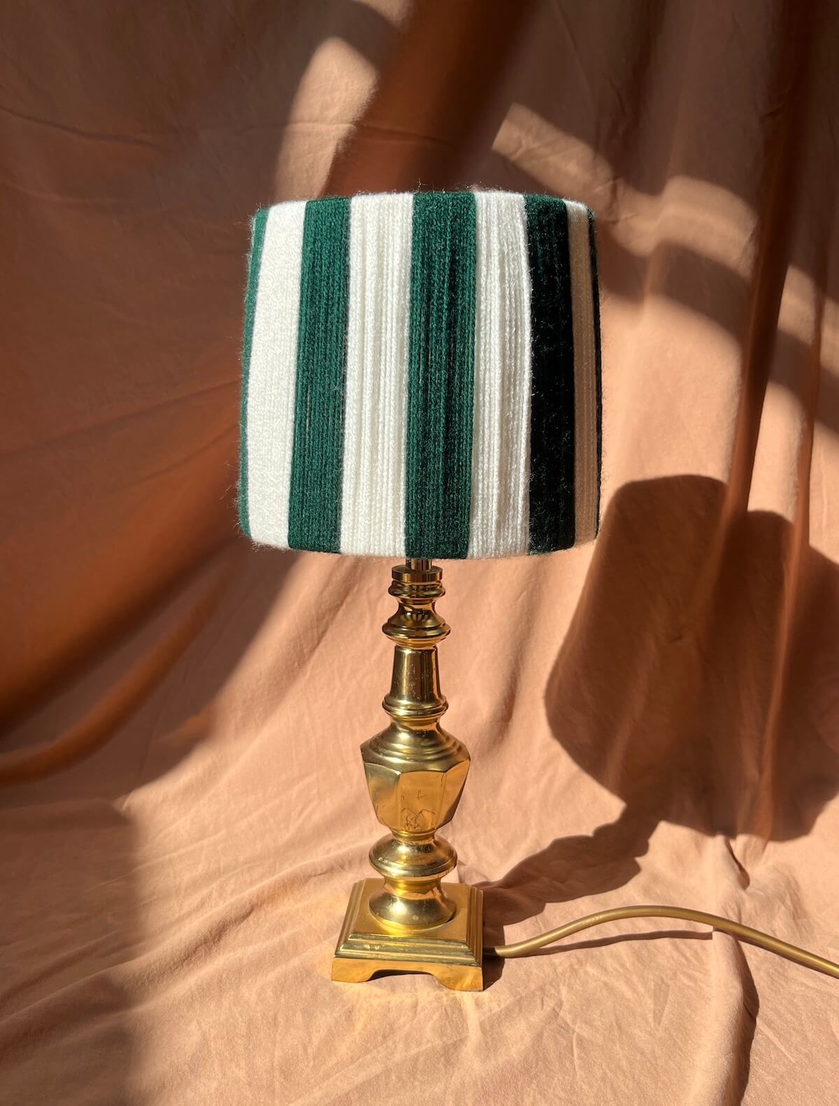 A Slice Shades table lamp with a green and white striped string shade and a polished gold brass base, set against a draped peach fabric background with dramatic shadows.