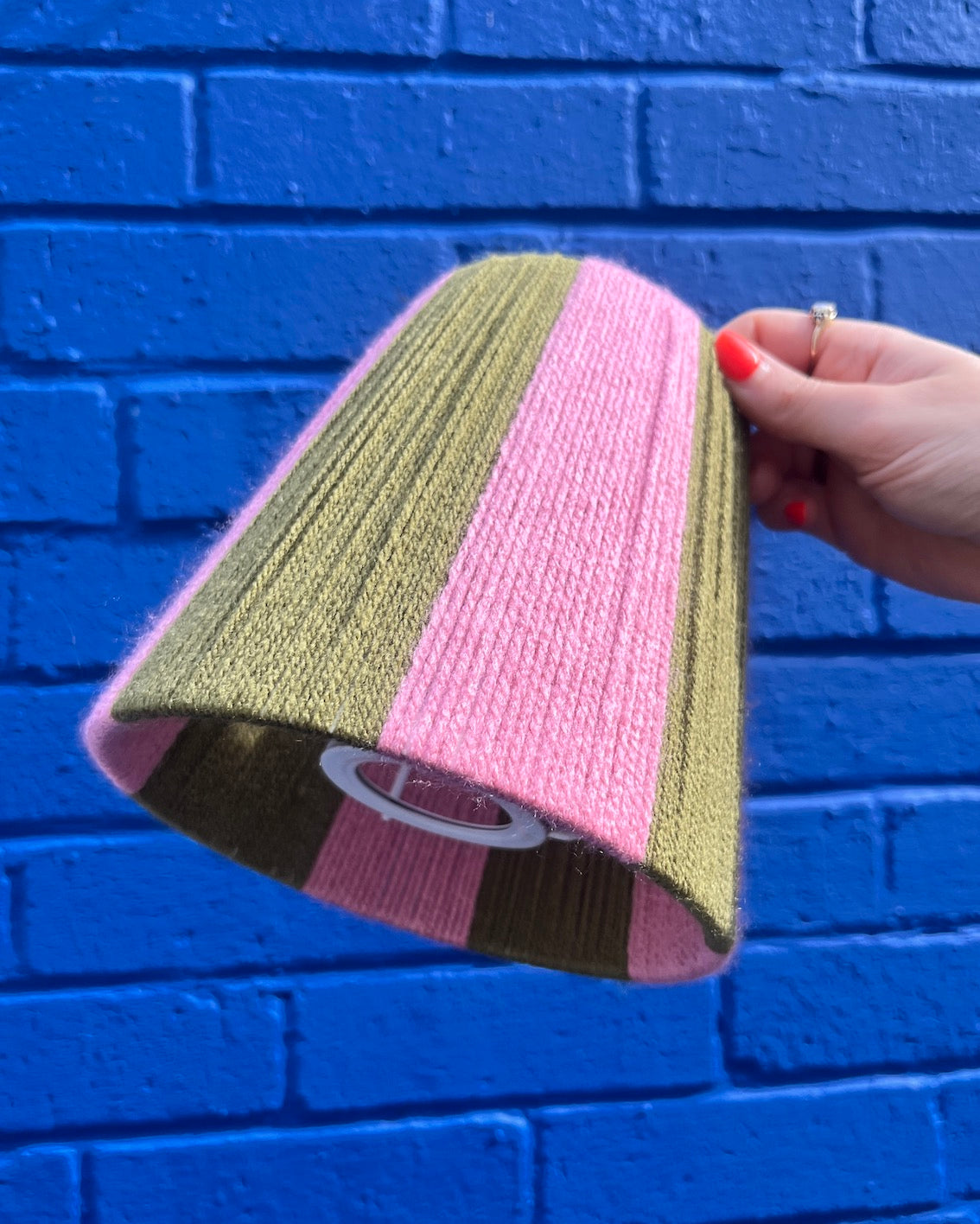 A hand holding a lice Shades yarn lampshade with vertical stripes of olive green and pink set against a bright blue painted brick wall.