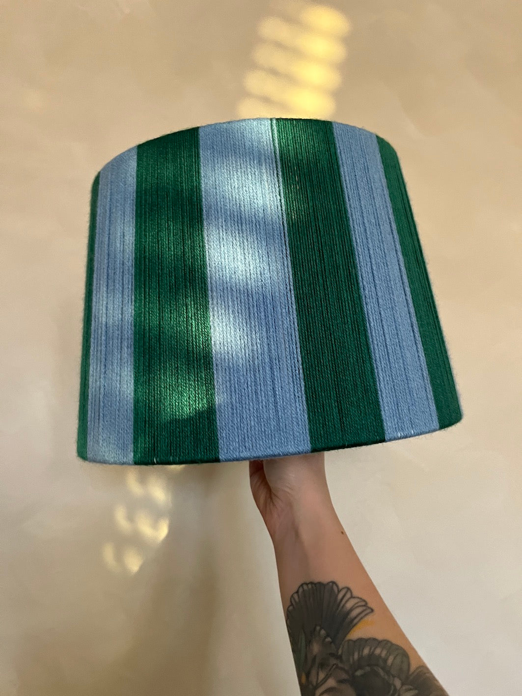 A hand with a tattoo holds up a Slice Shades handmade yarn drum lampshade featuring vertical stripes of forest green and light blue, set against a warm beige background with soft window light reflections.