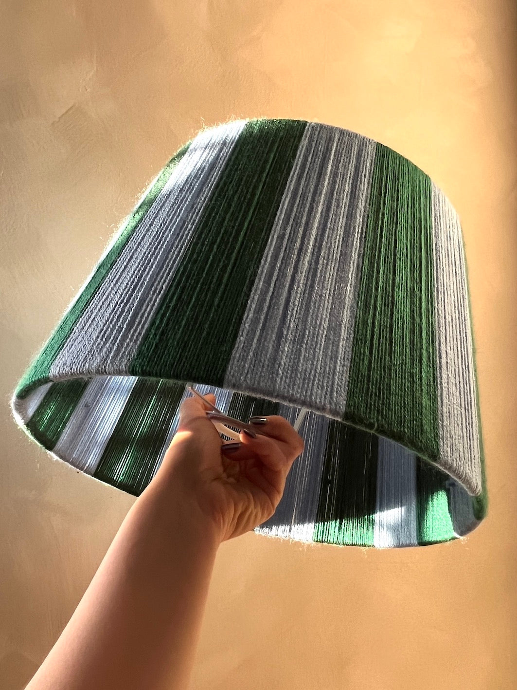 A hand holding a Slice Shades handmade yarn drum lampshade with vertical stripes of green, white, and gray, set against a warm, textured beige background.
