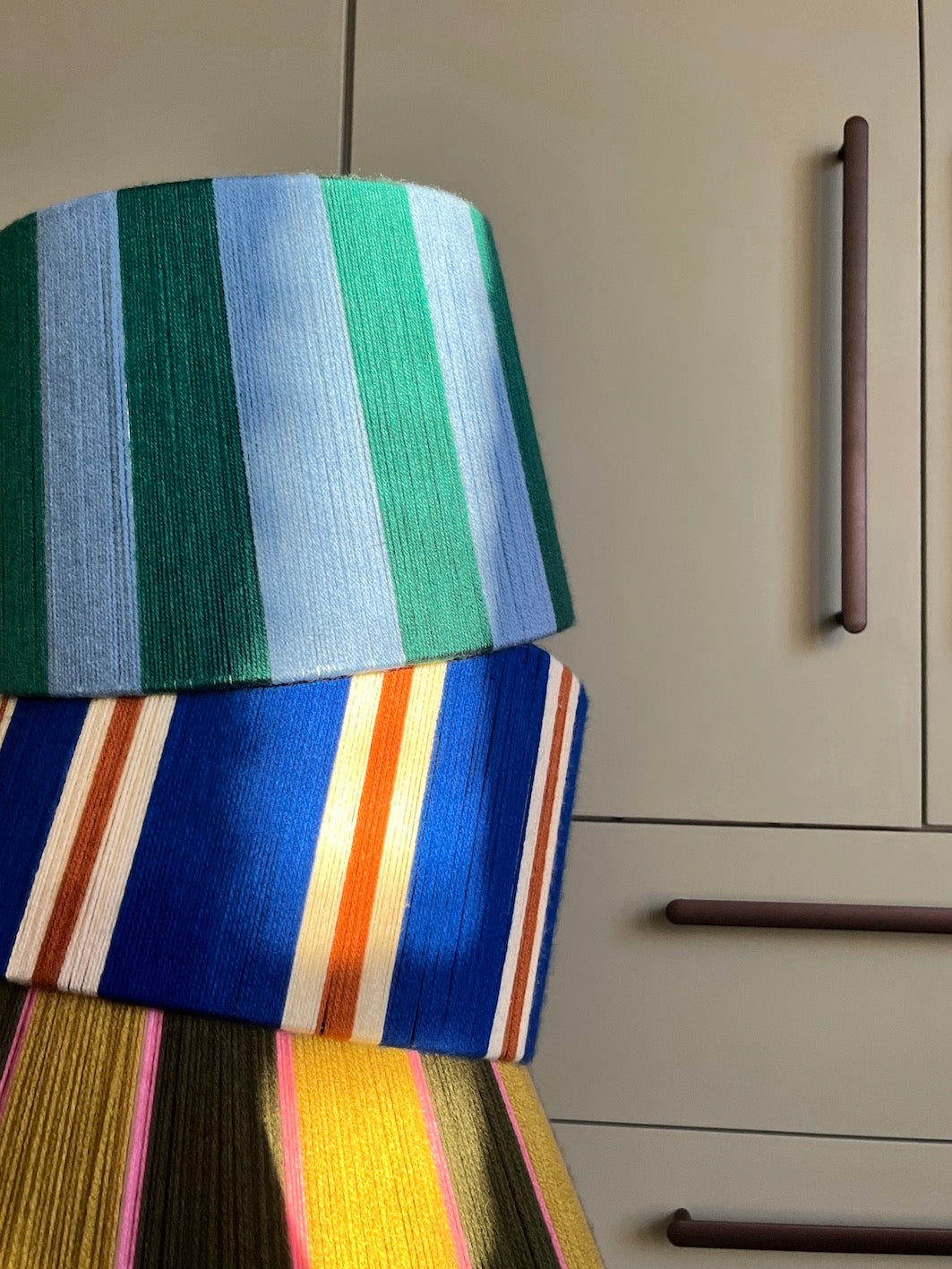 A close-up of Slice Shades handmade yarn drum lampshades with vertical stripes in green, blue, orange, cream, yellow, and pink, set against a beige cabinet background.