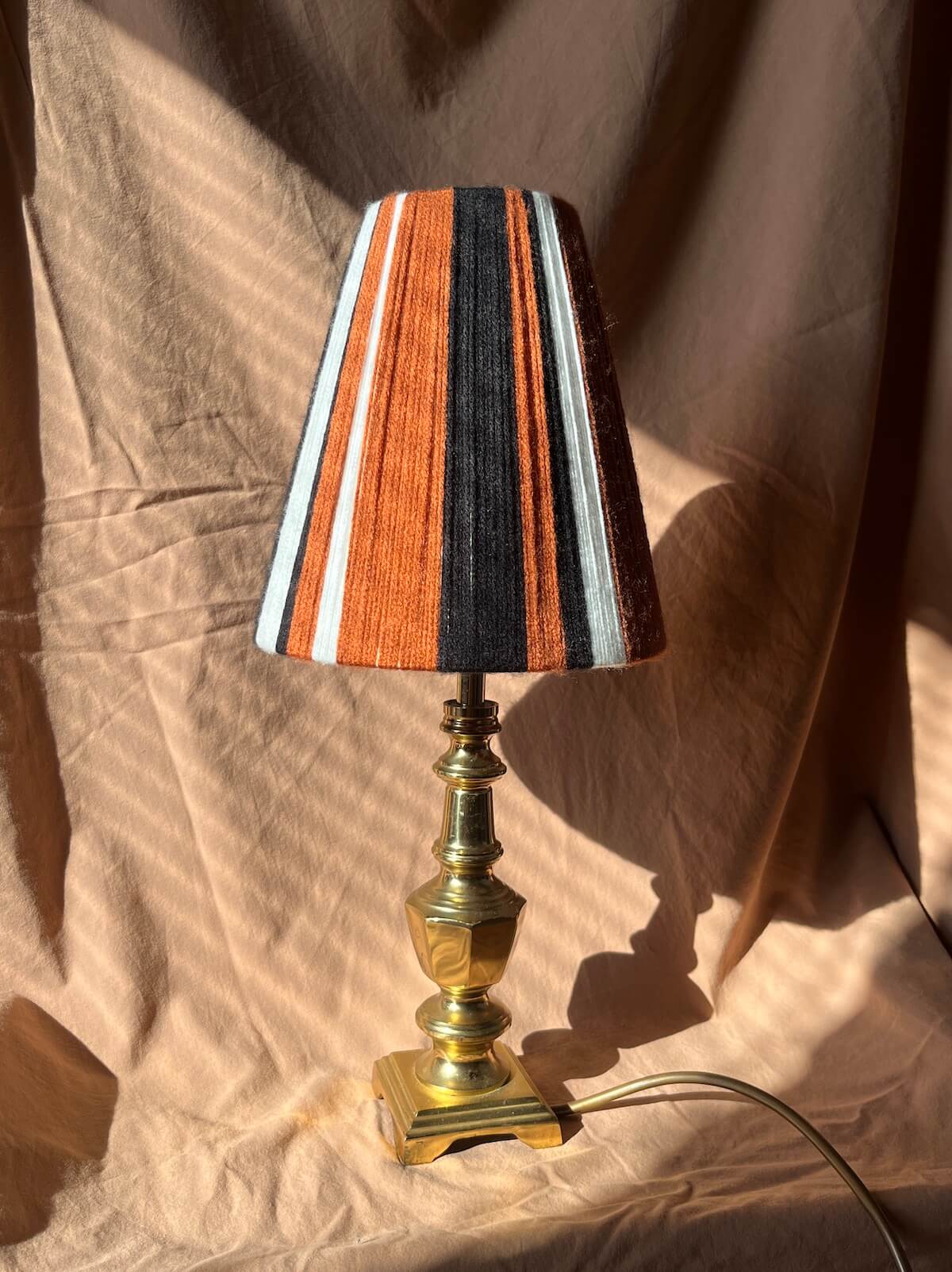 A Slice Shades table lamp with a striped yarn lampshade in rust, black, white, and brown, mounted on a polished brass base, set against a draped beige fabric backdrop.