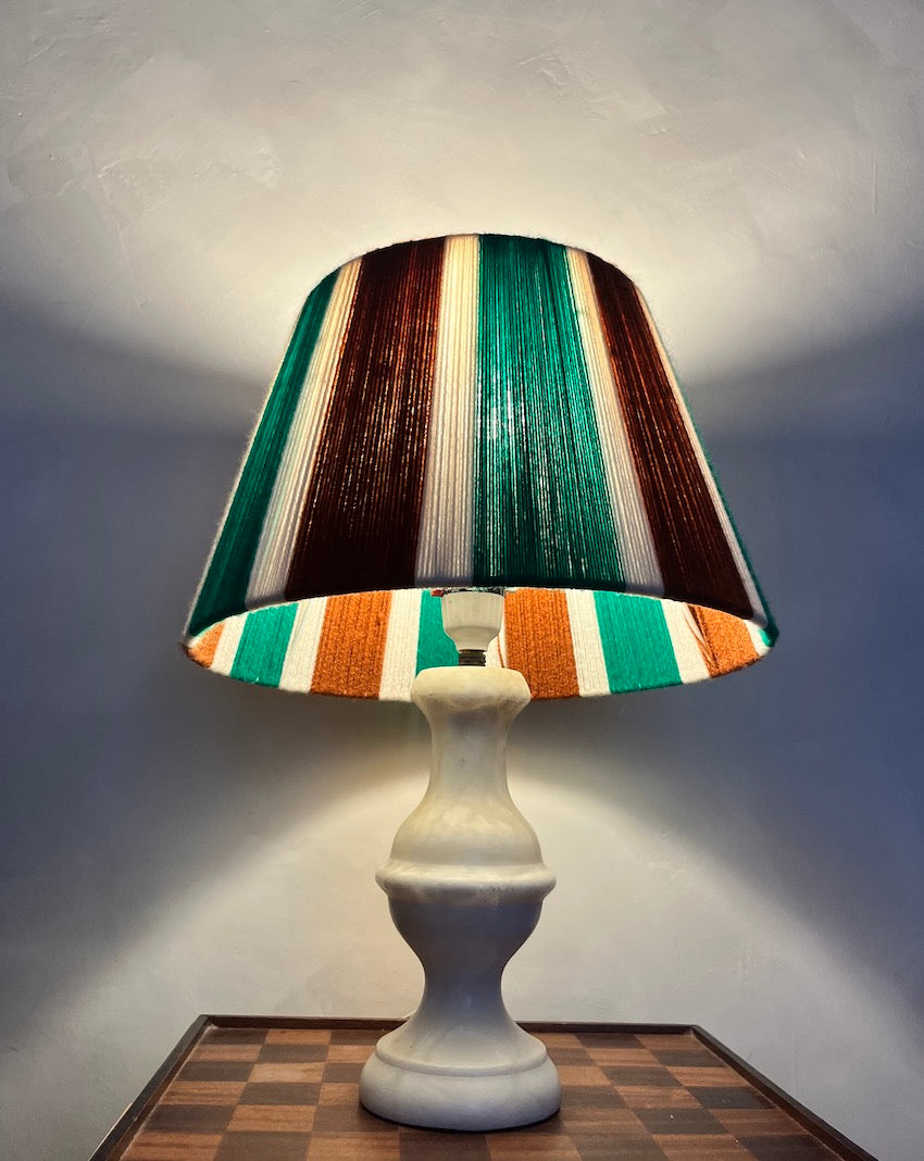 A table lamp with a sculptural white ceramic base and a Slice Shades yarn lampshade featuring vertical bands of green, brown, orange, and cream, shown lit against a gray wall.