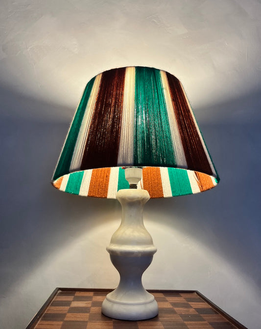 A table lamp with a sculptural white ceramic base and a Slice Shades yarn lampshade featuring vertical bands of green, brown, orange, and cream, shown lit against a gray wall.