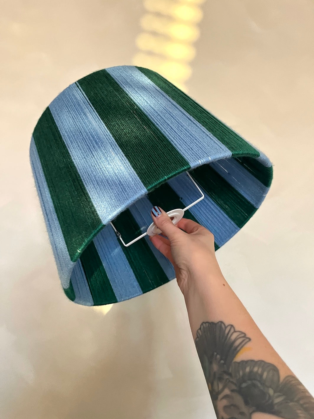 A hand with a tattoo holds Slice Shades handmade yarn drum lampshade with vertical stripes of dark green, light blue, and white. The lampshade is shown against a warm, beige tiled background.